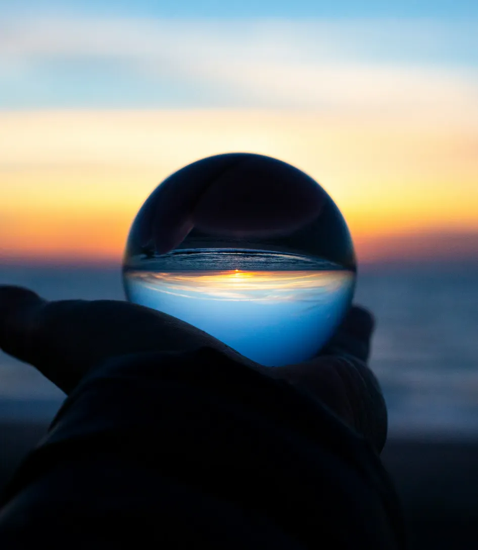 En hånd holder en glasskule som speiler en opp-ned solnedgang ved havet.