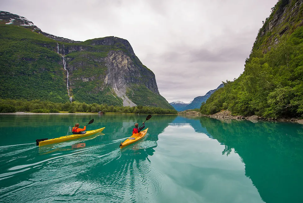 Zwei Personen paddeln im Kajak auf einem türkisfarbenen Fjordsee, umgeben von steilen Bergen und einem Wasserfall in Stryn, Norwegen.