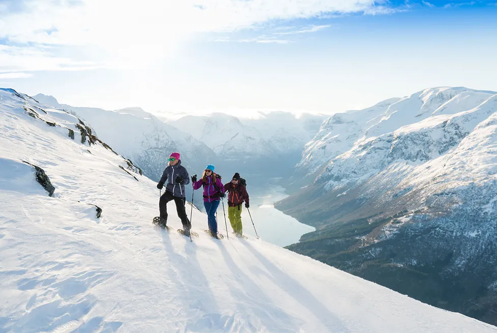 Drei Schneeschuhwanderer überqueren einen schneebedeckten Berghang über einem Fjord bei Stryn in Norwegen.