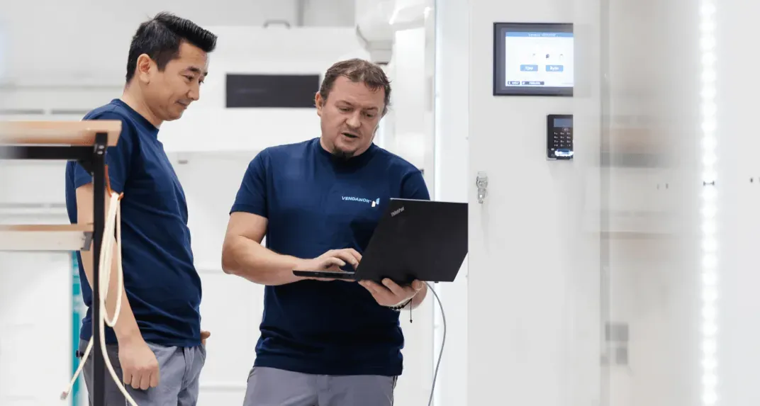 Two Vendanor technicians test a vending machine from a laptop beside its touchscreen and card terminal.