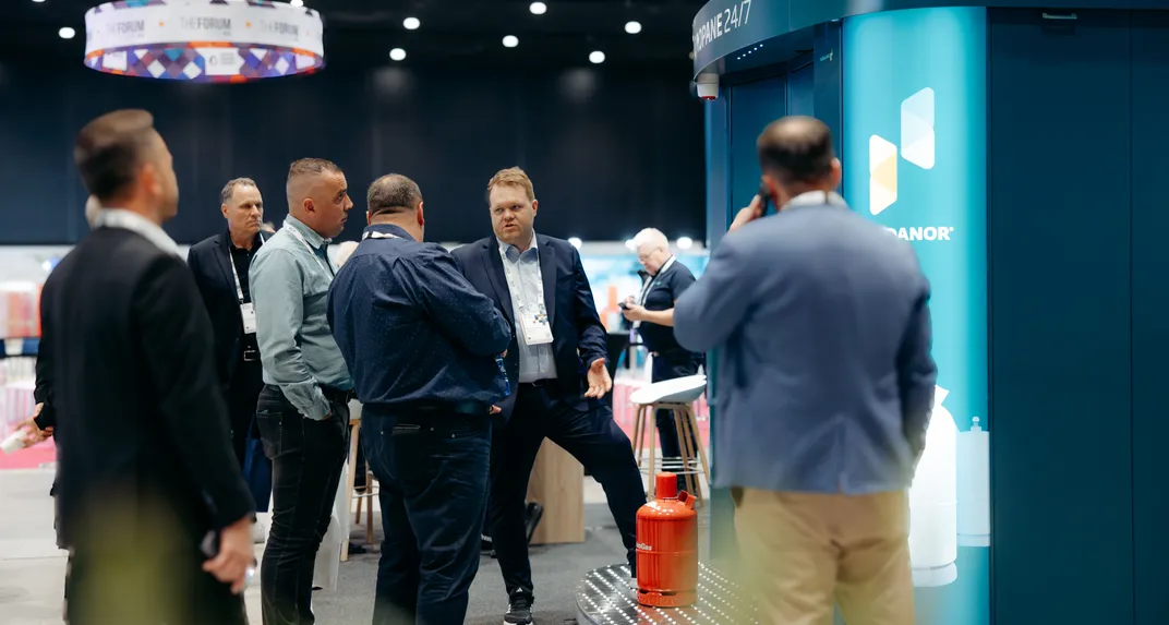 Visitors with lanyards talk beside a teal Vendanor Propane Vending Machine and a red cylinder at a trade fair.