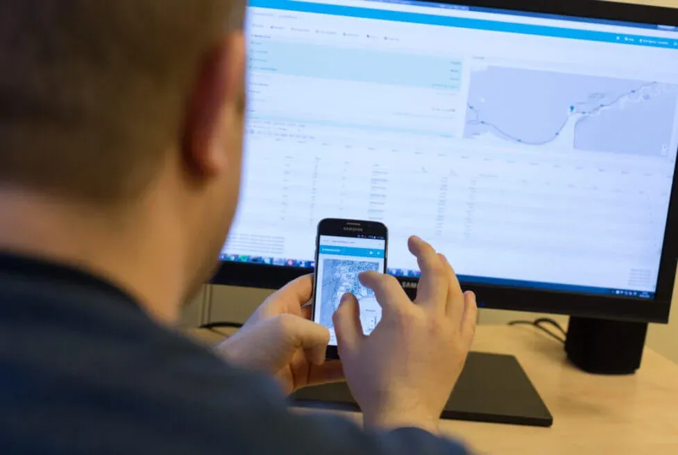 Eine Person fotografiert einen Telefonbildschirm, während sie das Vendanor Connect Management-Dashboard auf einem Monitor verwendet.