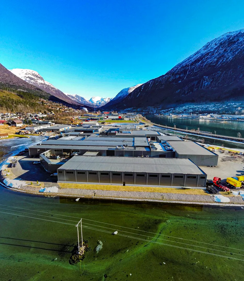 Flyfoto av Vendanor-fabrikken ved en fjord omgitt av snøkledde fjell.