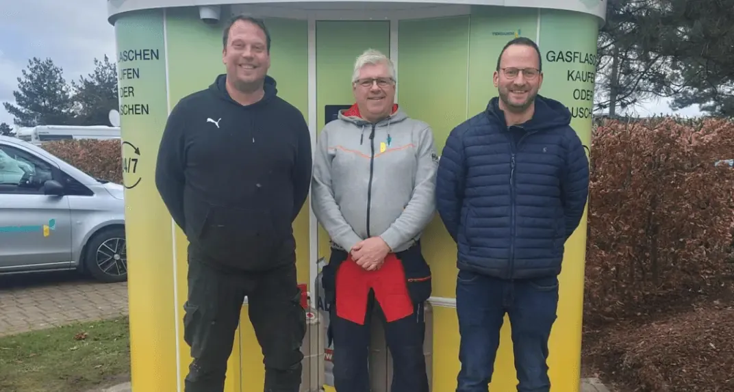 Three men standing in front of a green and yellow Vendanor Propane Vending Machine at Emsland camp.