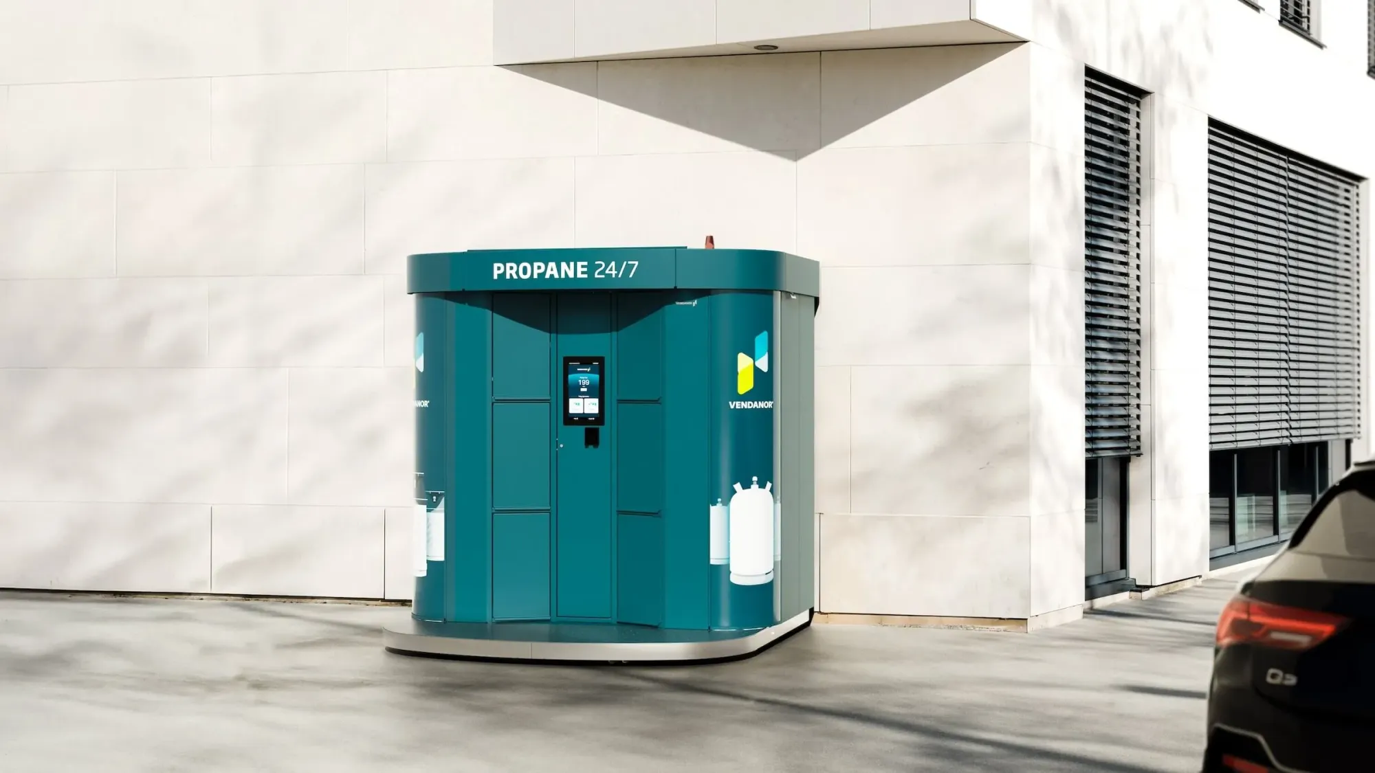 Ein blaugrüner Vendanor Gasflaschenautomat mit der Aufschrift Propane 24/7 steht vor einem modernen Gebäude neben einem geparkten Auto.