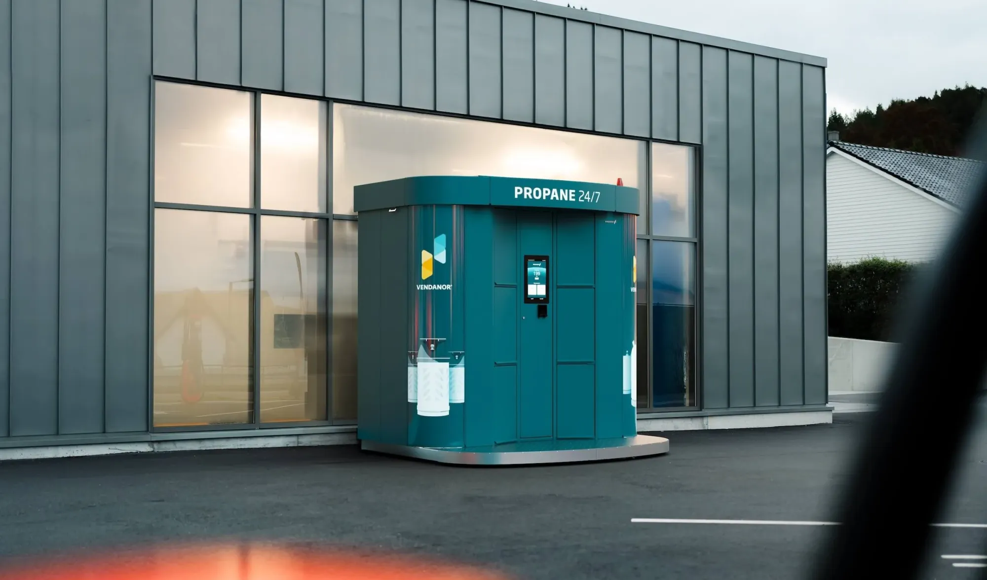 Ein blaugrüner Vendanor Gasflaschenautomat mit der Aufschrift Propane 24/7 steht neben einem grauen Industriegebäude, gesehen durch ein Autofenster.