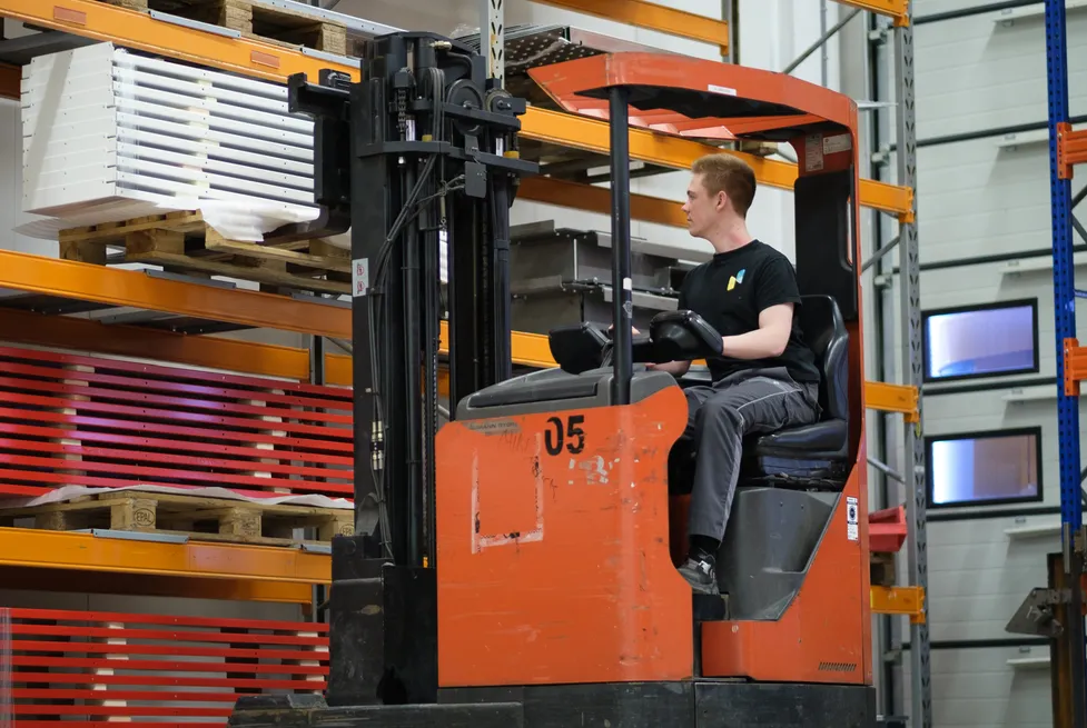A Vendanor worker operates an orange reach-truck forklift among warehouse storage racks.