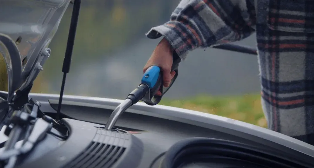 A person in a plaid shirt fills a car's washer fluid reservoir with the nozzle of a Vendanor Washer Fluid Dispenser.
